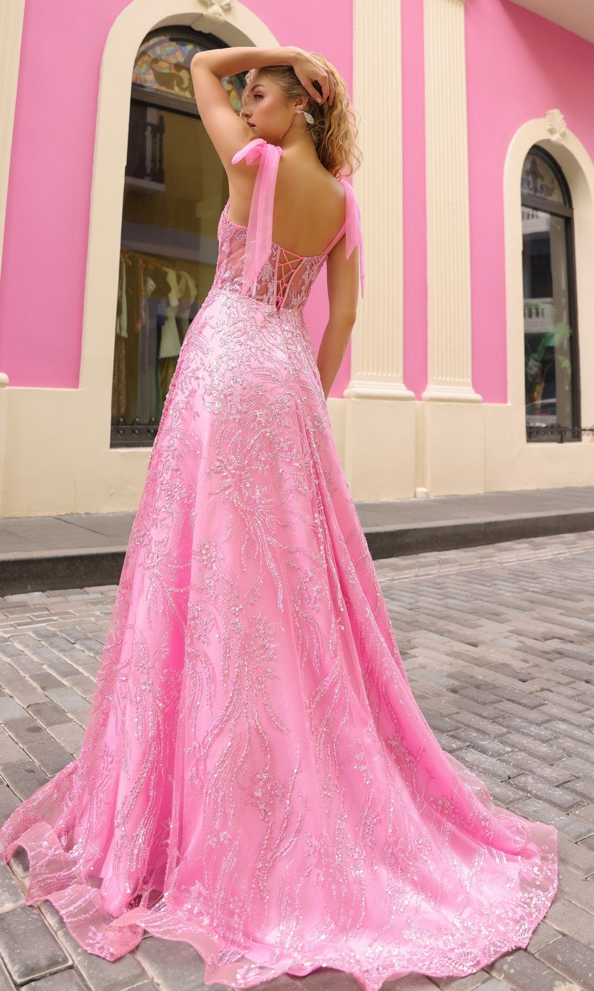 A woman poses outdoors on a cobblestone street in front of a pink and cream building, wearing the Sequin Long Prom Ball Gown with Shoulder Bows T1336.